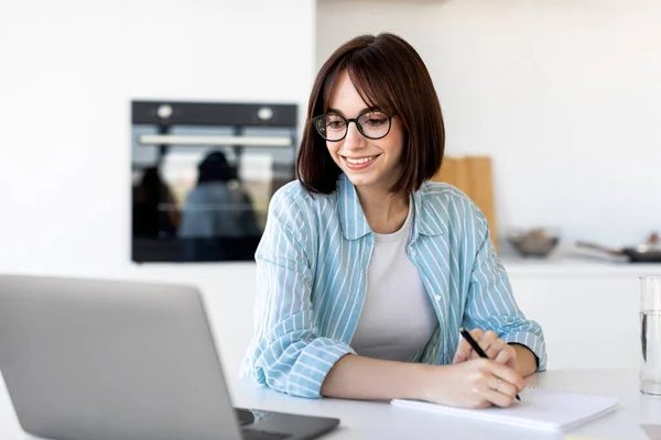 Senhora freelancer animado trabalhando com laptop no interior da cozinha e tomar notas, escrevendo informações — Fotografia de Stock