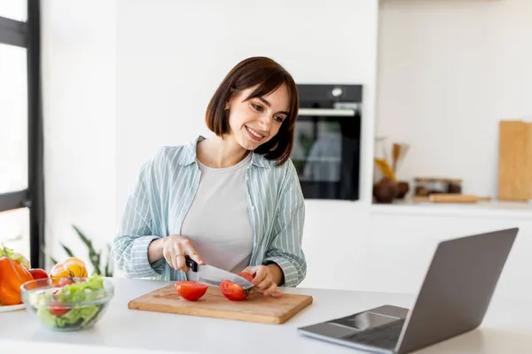 Junge Frau kocht frischen Biosalat, schneidet Tomaten und schaut auf Laptop auf Arbeitsplatte, probiert neues Rezept aus — Stockfoto