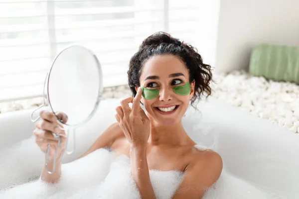 Beauty day and home skin care. Lovely young female applying patches under eyes, looking in mirror, lying in foamy bath