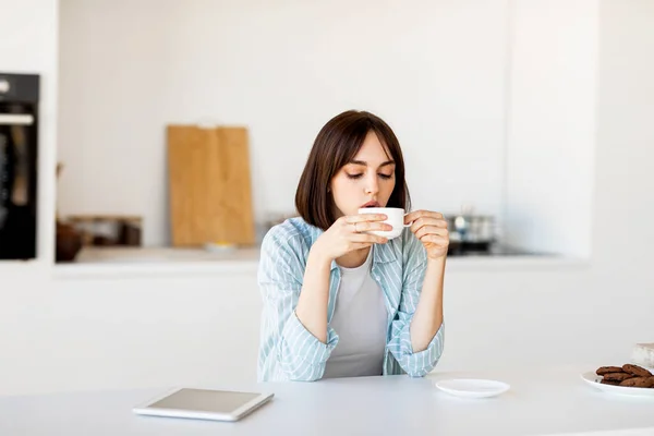 Jonge ontspannen dame zit in de moderne keuken, het drinken van koffie, genieten van warme drank in de ochtend — Stockfoto