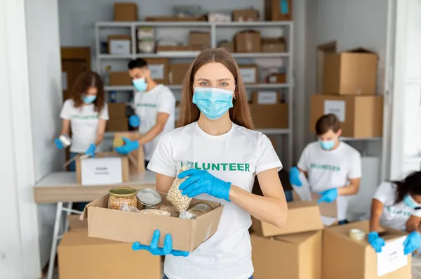 Jovem voluntária vestindo uniforme voluntário e máscara médica, segurando caixa com alimentos enlatados e grãos — Fotografia de Stock