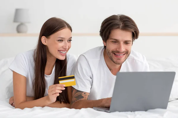 Boldog millenniumi kaukázusi nő és a srác fekszik az ágyon fizet a vásárlásért, rendelés laptop és hitelkártya — Stock Fotó