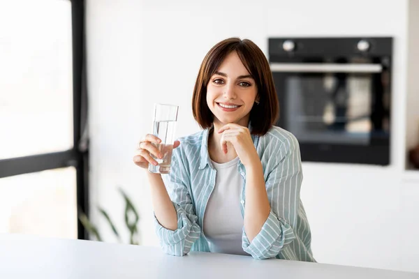 Porträtt av ung dam dricksvatten från glas, sitter i köket interiör, ta hand om kroppen hydratisering — Stockfoto