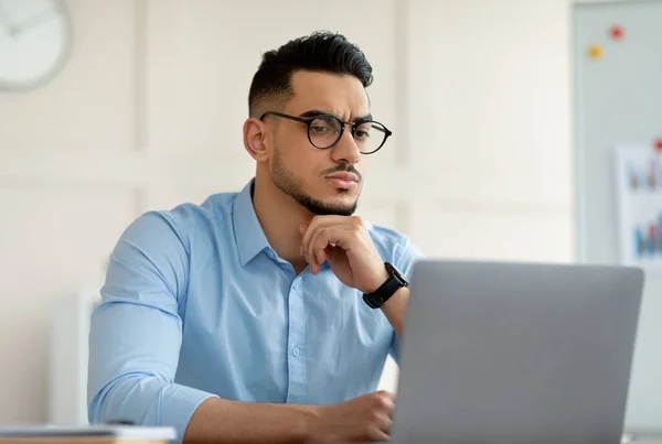 Unruhiger arabischer Büroangestellter sitzt am Tisch, schaut auf den Laptop-Bildschirm und denkt über geschäftliche Probleme im Büro nach — Stockfoto