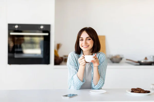 Portret van een vrolijke freelancer dame die geniet van een kopje hete koffie in de keuken en glimlacht naar de camera, vrije ruimte — Stockfoto