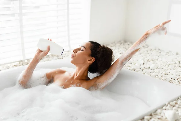 Charmante jonge vrouw nemen schuimige bad, met behulp van shampoo fles als mic, zingen populaire lied, plezier hebben thuis — Stockfoto