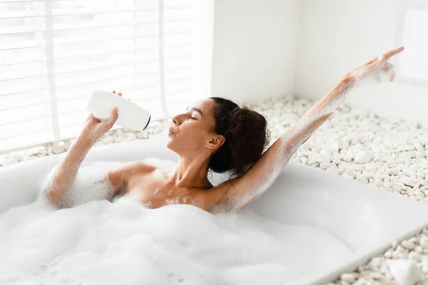 Encantadora joven tomando un baño espumoso, usando una botella de champú como micrófono, cantando una canción popular, divirtiéndose en casa —  Fotos de Stock