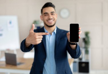 Genç Arap işadamı modern ofiste boş ekranlı akıllı telefonu işaret ediyor, mobil uygulama veya web sitesi için model