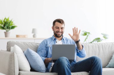 Orta yaşlı mutlu Avrupalı adamın oturma odasında el sallarken görüntüsü var.