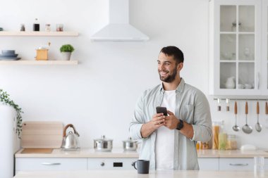 Akıllı telefonlu ve bir fincan kahveli mutlu beyaz adam mutfağın içindeki boşluğa bakıyor.