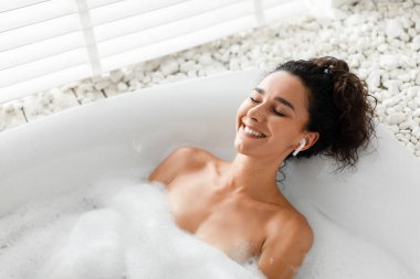Sakin gülümseyen genç kadın köpük banyosunda gözleri kapalı dinleniyor, kulaklıkla müzik veya sesli meditasyon dinliyor.