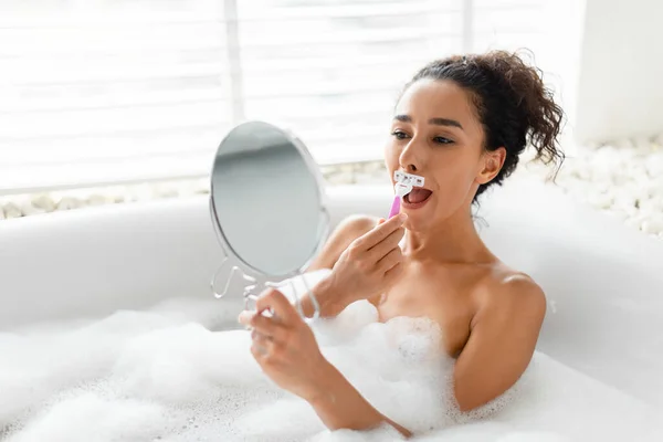 Jolie jeune femme rasant sa lèvre supérieure, enlever la moustache indésirable dans le bain moussant devant le miroir à la maison — Photo