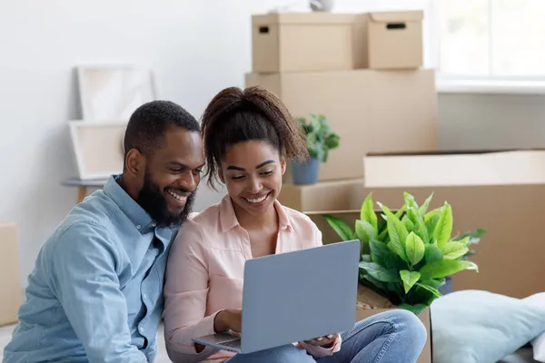 Χαμογελώντας νεαρός μαύρος άνδρας και η κυρία σε νέο διαμέρισμα μεταξύ κουτιά, σχεδιασμό εσωτερικό, ζευγάρι που χρησιμοποιούν φορητό υπολογιστή — Φωτογραφία Αρχείου