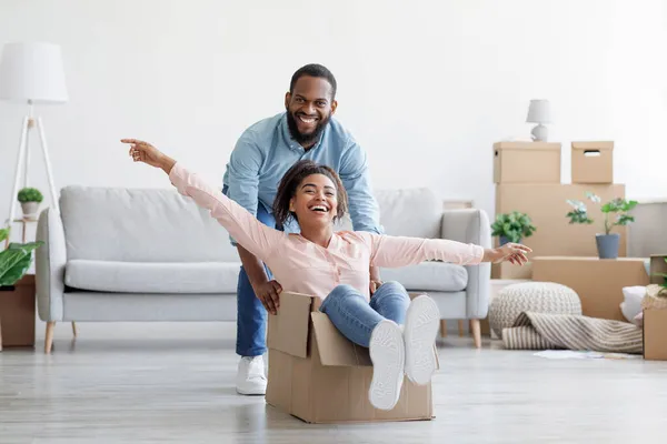 Satisfeito rindo jovem afro-americano marido empurrando caixa com mulher animada em novo apartamento na sala de estar — Fotografia de Stock