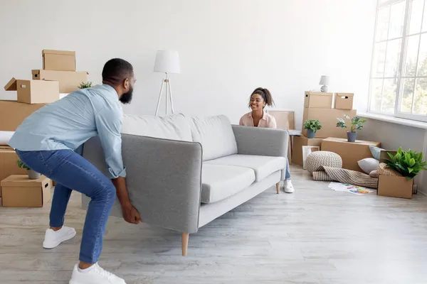 Alegre milenar afro-americano marido e mulher carregam sofá em novo apartamento com caixas na sala de estar — Fotografia de Stock