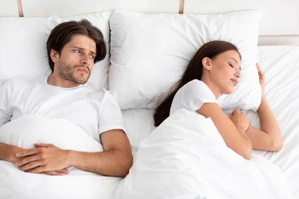 Descontente triste caucasiano jovem não dorme, encontra-se na cama, olha para senhora, mulher ronca no quarto branco — Fotografia de Stock