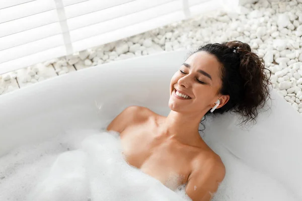 Senyum tenang wanita muda santai di bubble bath dengan mata tertutup, mendengarkan musik atau audio meditasi di earphone — Stok Foto