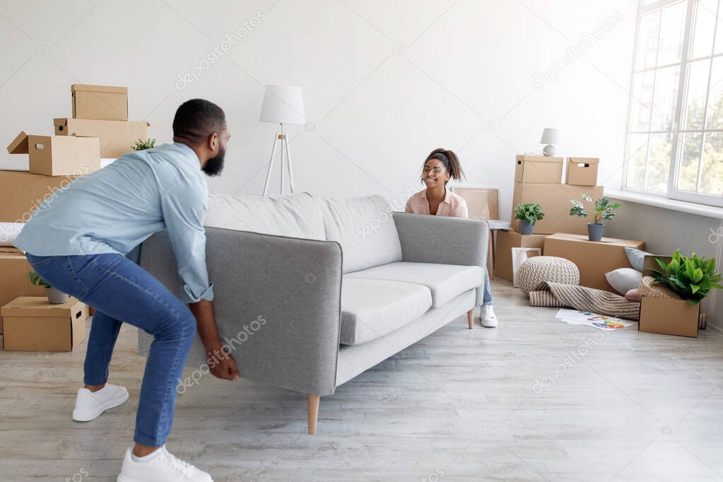 Cheerful millennial african american husband and wife carry sofa in new apartment with boxes in living room