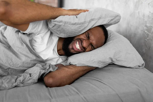 Hombre negro estresado cubriendo las orejas acostado en la cama — Foto de Stock