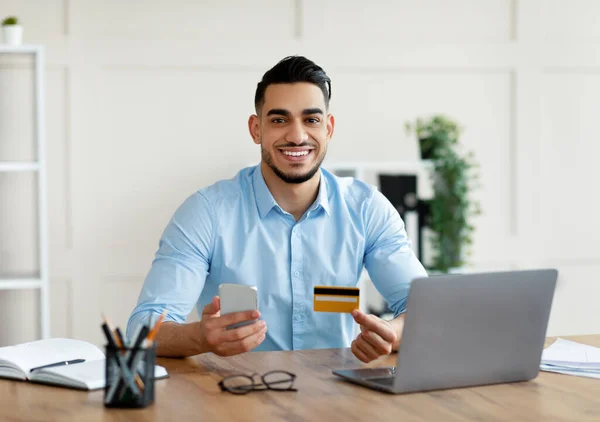 Wesoły młody Arab posiadający kartę kredytową i komórkę, dokonujący zdalnej płatności, zamawiający towary w biurze — Zdjęcie stockowe