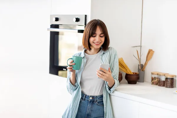 Chat in de ochtend, nieuws en ontbijt thuis. Gelukkig jonge dame drinken van koffie en kijken naar smartphone in de keuken — Stockfoto