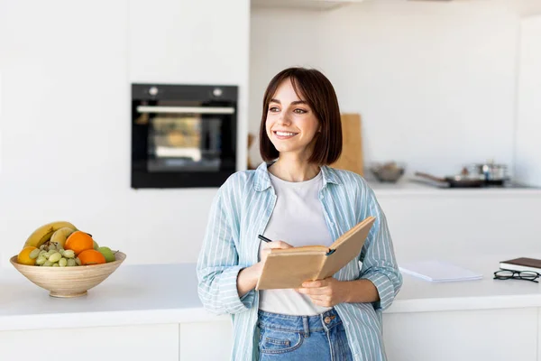 Hübsche Frau macht Notizen, hält Notizblock und Stift in der Hand, steht in der Küche und schaut zur Seite, Kopierraum — Stockfoto