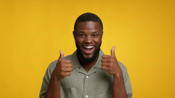 Me gusta. Chico negro lleno de alegría mostrando pulgares hacia arriba en la cámara, alegre juguetón afroamericano bailando mientras muestra señal de aprobación, posando sobre fondo de estudio amarillo, grabación en cámara lenta — Vídeo de stock