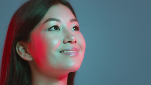 Молодая красивая азиатка смотрит на свободное пространство и искренне улыбается, выражая вдохновение, синий студийный фон — стоковое видео