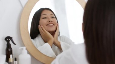 Başarılı yerli güzellik hizmetleri. Bornoz giymiş genç ve mutlu Asyalı bayan aynaya bakıyor ve gülümsüyor.