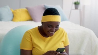 Evde akıllı telefon kullanırken gülen pozitif siyah kavisli kadın.