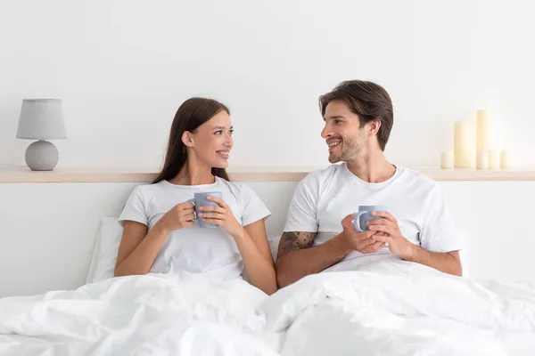 Alegre feliz jovem europeu marido e mulher olham um para o outro, segurar copos na cama no interior do quarto — Fotografia de Stock