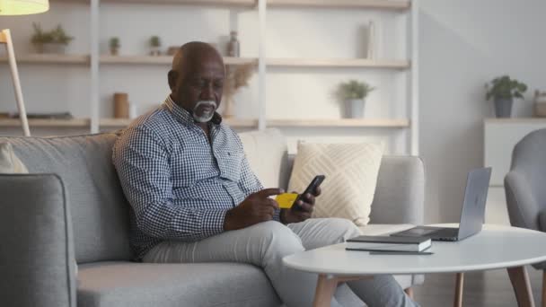 Banca móvil. Positivo hombre afroamericano senior compras en línea con tarjeta de crédito y aplicación móvil en casa — Vídeos de Stock