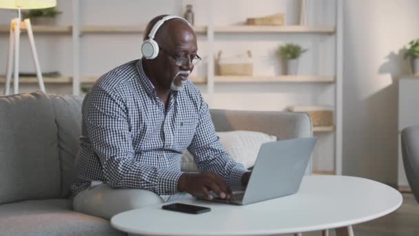 Positivo uomo afroamericano anziano alla moda ascoltando musica in cuffie wireless mentre si lavora su laptop a casa — Video Stock