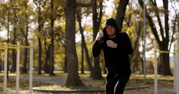 Aktív életmód és sport. Fiatal sportos srác fekete kapucnis edzőcipőben boksz ütések a szabadban, edzés az őszi parkban — Stock videók