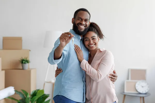 Χαρούμενη χαμογελαστή νεαρή αφροαμερικάνικη αγαπημένη οικογένεια που αγκαλιάζεται σε νέο σπίτι ανάμεσα σε κουτιά και δείχνει τα κλειδιά του σπιτιού. — Φωτογραφία Αρχείου