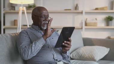 Uzaktan iletişim. Afrika kökenli Amerikalı büyükbaba ailesiyle görüntülü konuşma yapıyor, evdeki dijital tablete el sallıyor.