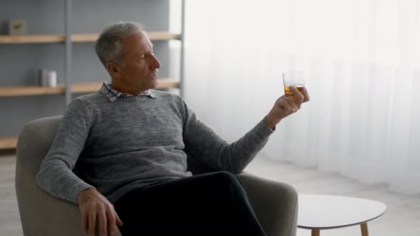 Senior man die whisky drinkt terwijl hij thuis ontspant in een fauteuil — Stockvideo