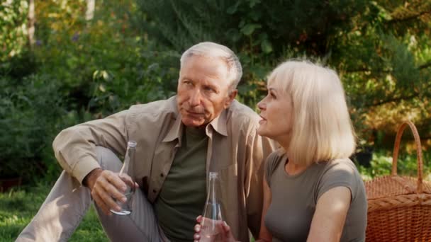 Ältere Ehepartner entspannen sich draußen im Garten, trinken Wasser aus Flaschen und plaudern, glückliches reifes Paar, das draußen picknickt, die gemeinsame Zeit in der Natur genießen, Zeitlupe filmen — Stockvideo