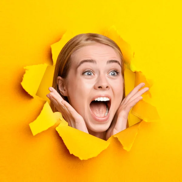 Blondynka krzycząca OMG, zszokowana ogromną sprzedażą lub rabatem, zaglądająca przez dziurę w podartym pomarańczowym papierze — Zdjęcie stockowe