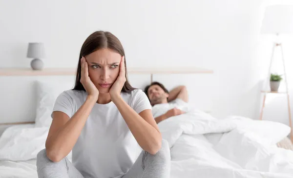 Offeso triste sconvolto bella giovane donna caucasica si siede sul letto mentre il marito dorme in camera da letto interno — Foto Stock