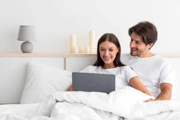 Souriant attrayant jeune mari caucasien avec chaume câlins femme assise sur le lit, utiliser un ordinateur portable pour surfer sur Internet — Photo