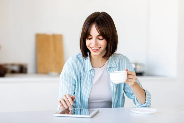 Boldog hölgy használ digitális tabletta, miközben élvezi a reggeli kávét a konyhában otthon, ül az asztalnál — Stock Fotó