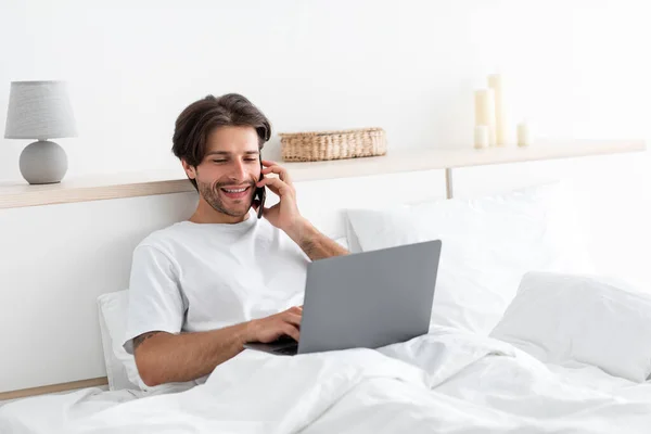 Wesoły, atrakcyjny, millenijny Europejczyk pracuje na laptopie, rozmawia przez telefon z klientem na łóżku — Zdjęcie stockowe