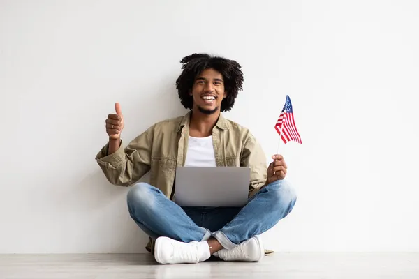 Glücklicher junger schwarzer Kerl sitzt auf dem Boden mit Laptop und amerikanischer Flagge — Stockfoto