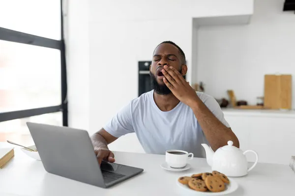 Hombre negro cansado bostezando usando el ordenador portátil sentado en la mesa de la cocina —  Fotos de Stock