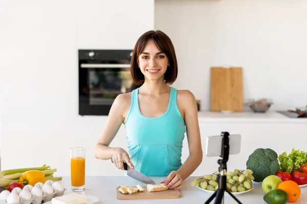 Aufgeregte schlanke Dame, die Obstsalat zubereitet, Video für ihr Vlog auf dem Smartphone aufnimmt, in der Küche steht — Stockfoto