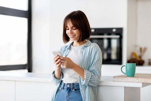 On-line objednávání jídla. Nadšený mladá dáma psaní na smartphone v moderním kuchyňském interiéru, volné místo — Stock fotografie