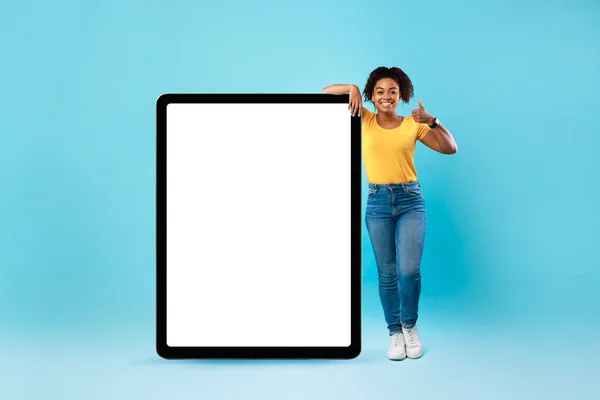 Senhora preta feliz de pé perto do computador tablet enorme com tela vazia, mostrando polegar para cima, recomendando aplicativo ou site, mockup — Fotografia de Stock