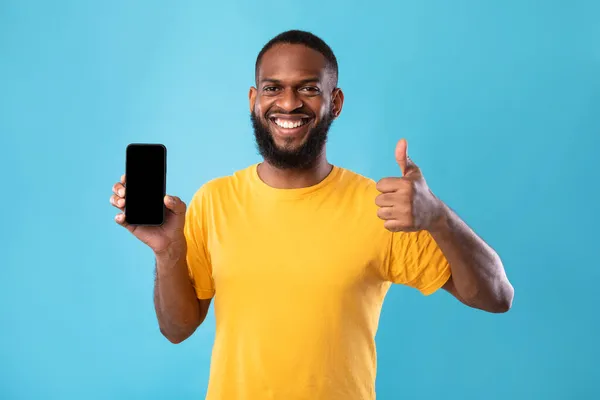 Jovem negro mostrando smartphone com tela em branco, gesticulando polegar para cima, recomendando novo aplicativo ou site, mockup — Fotografia de Stock