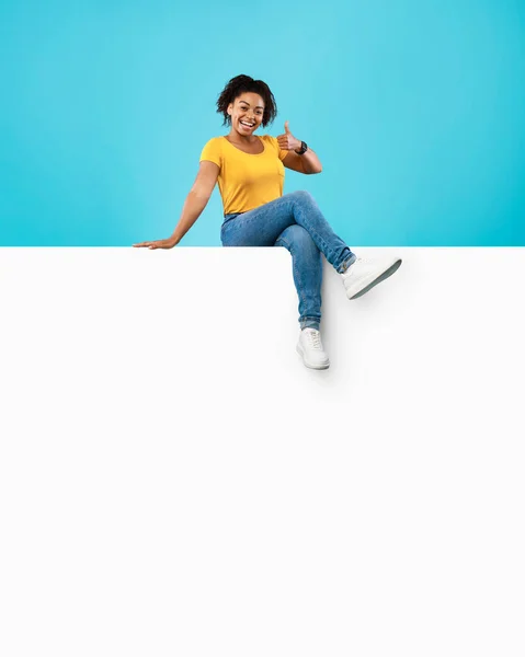 Sonriente mujer afroamericana sentada en un póster vacío, mostrando un gesto de pulgar hacia arriba en el fondo del estudio azul, maqueta —  Fotos de Stock